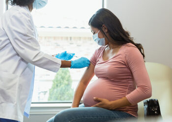 Wearing protective masks because of COVID-19, a mid adult pregnant woman watches as the female technician administers a booster shot.