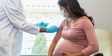 Wearing protective masks because of COVID-19, a mid adult pregnant woman watches as the female technician administers a booster shot.