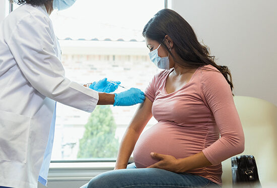 Wearing protective masks because of COVID-19, a mid adult pregnant woman watches as the female technician administers a booster shot.