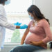 Wearing protective masks because of COVID-19, a mid adult pregnant woman watches as the female technician administers a booster shot.